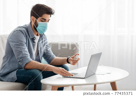 Gadgets sterilization. Arab man freelancer in face mask cleaning laptop keyboard with spray and napkin, sitting on sofa Gadgets sterilization. Arab man freelancer in face mask cleaning laptop keyboard with spray and napkin, sitting on sofa 77435371