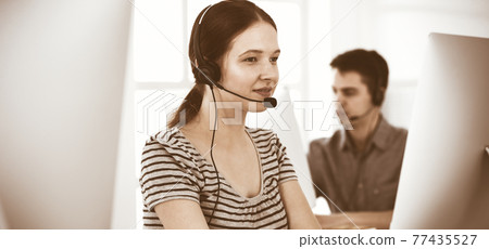Casual dressed young woman using headset and computer while talking with customers online. Group of operators at work. Call center, business concept Casual dressed young woman using headset and computer while talking with customers online. Group of operators at work. Call center, business concept 77435527