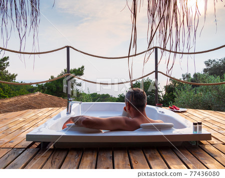 A handsome adult man lies in a hot tub at a luxurious exotic spa resort hotel balcony with sea and sunset view. 77436080