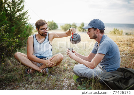 Outdoors image of young explorer man drinking hot beverage in mountains, sitting and relaxing after trekking. Outdoors image of young explorer man drinking hot beverage in mountains, sitting and relaxing after trekking. 77436175