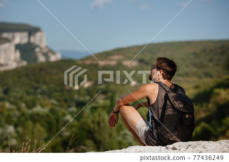 Young male hiker with backpack relaxing on top of a mountain during calm summer sunset Young male hiker with backpack relaxing on top of a mountain during calm summer sunset 77436249