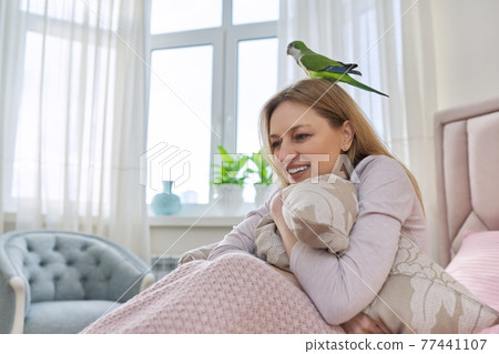 Happy laughing mature woman and green parrot at home in bed Happy laughing mature woman and green parrot at home in bed 77441107