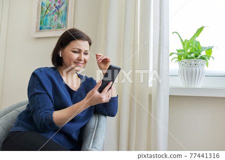 Smiling middle-aged woman in wireless headphones looking at smartphone screen 77441316