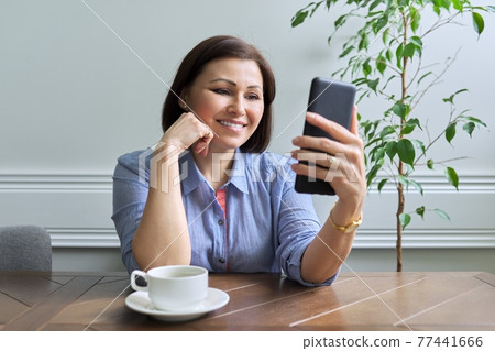 Mature woman with smartphone sitting at home smiling looking at phone screen Mature woman with smartphone sitting at home smiling looking at phone screen 77441666