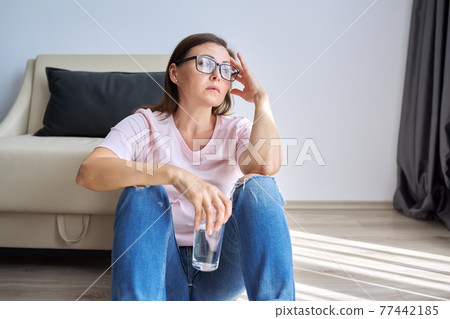 Sad unhappy mature woman sitting at home on floor with glass of water Sad unhappy mature woman sitting at home on floor with glass of water 77442185