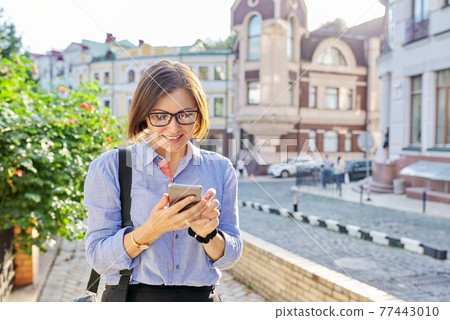 Mature business woman typing on smartphone walking along city street. 77443010