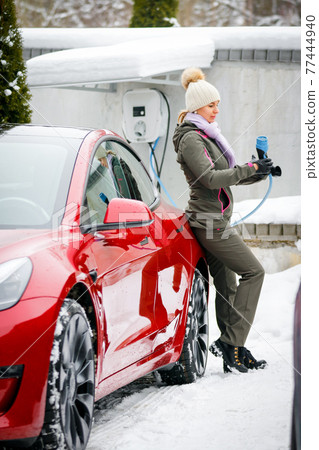 Stylish girl stands near her red electric car and waits for the car to charge. Connecting the plug of the electric vehicle charger. Stylish girl stands near her red electric car and waits for the car to charge. Connecting the plug of the electric vehicle charger. 77444940