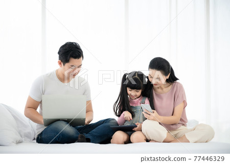 Father and mother with daughter relaxing and using laptop and smartphone on bed 77446329