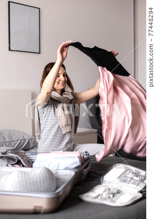 Happy woman putting clothes and accessories into suitcase getting ready to travel vacation on bed 77448294