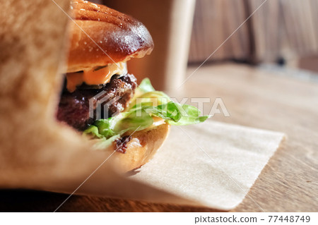 Burger in paper packaging close-up. Food in a fast food restaurant. Macro photo of burger ingredients with meat and vegetables Burger in paper packaging close-up. Food in a fast food restaurant. Macro photo of burger ingredients with meat and vegetables 77448749