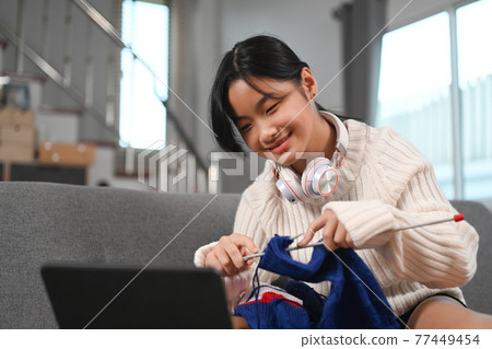 Happy young girl enjoying knitting and watching something on digital at home. Happy young girl enjoying knitting and watching something on digital at home. 77449454