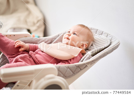 Young mother feeding her baby daughter with fruit puree. The baby is 7 months old, lying in a special high chair for babies. Young mother feeding her baby daughter with fruit puree. The baby is 7 months old, lying in a special high chair for babies. 77449584