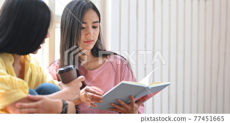Photo of young sisters relaxing/reading a notebook while sitting together at the wooden floor over comfortable living room windows as background. Photo of young sisters relaxing/reading a notebook while sitting together at the wooden floor over comfortable living room windows as background. 77451665