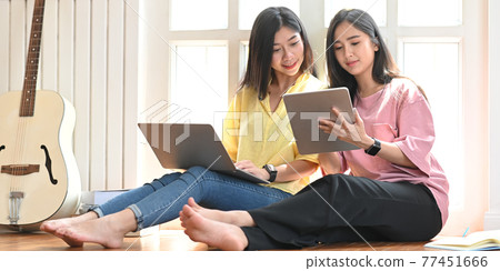 Young sisters holding and looking at computer tablet and laptop in hands while sitting together at the wooden floor over comfortable music studio windows as background. Young sisters holding and looking at computer tablet and laptop in hands while sitting together at the wooden floor over comfortable music studio windows as background. 77451666