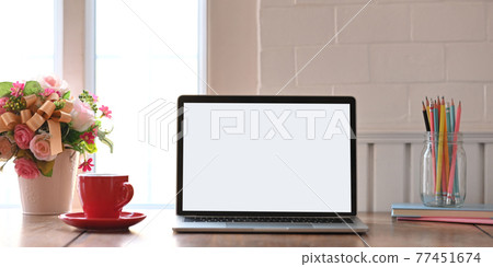Photo of computer laptop with white blank screen putting on wooden working desk and surrounded by notebook, bunch of flowers, cup and pencil holder over comfortable living room windows as background. 77451674