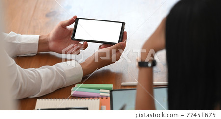 Photo of young businessman showing a white blank screen smartphone in his hands to colleague while sitting together at the wooden working table over orderly workspace as background. Photo of young businessman showing a white blank screen smartphone in his hands to colleague while sitting together at the wooden working table over orderly workspace as background. 77451675