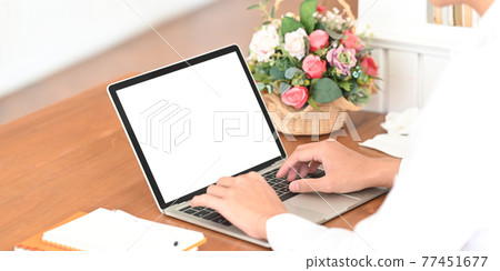 Cropped image of smart man's hands typing on computer laptop with white blank screen that putting on wooden working desk and surrounded by bunch of flowers in wicker basket, notebook and paperwork. Cropped image of smart man's hands typing on computer laptop with white blank screen that putting on wooden working desk and surrounded by bunch of flowers in wicker basket, notebook and paperwork. 77451677