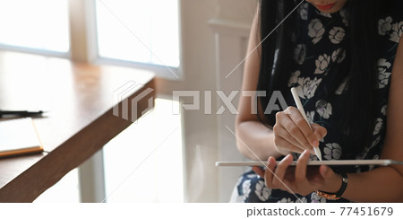 Cropped image of beautiful woman holding a computer tablet and stylus pen while sitting at the wooden working desk over comfortable living room as background. Cropped image of beautiful woman holding a computer tablet and stylus pen while sitting at the wooden working desk over comfortable living room as background. 77451679