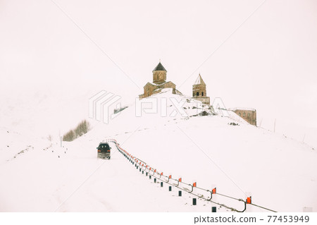 Stepantsminda, Gergeti, Georgia. Car Goes To Famous Ancient Gergeti Trinity Tsminda Sameba Church In Beautiful Georgian Winter Nature Landscape Stepantsminda, Gergeti, Georgia. Car Goes To Famous Ancient Gergeti Trinity Tsminda Sameba Church In Beautiful Georgian Winter Nature Landscape 77453949