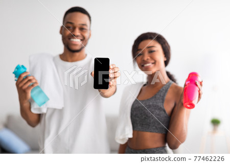 Young athletic black couple holding bottles of water and showing smartphone with empty screen at home, mockup 77462226