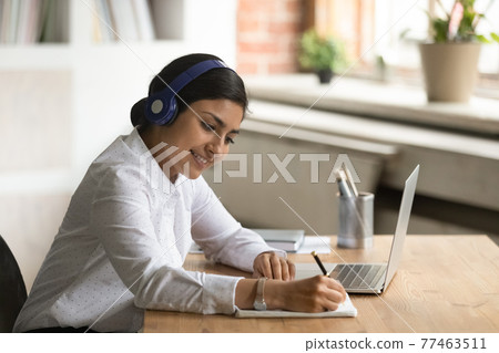 Smiling interested Indian woman in headphones taking notes, using laptop 77463511