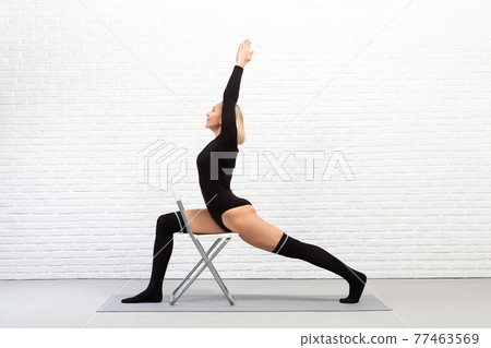 Iyengar yoga indoor with a chair. Sexy fit caucasian woman in black tight-fitting body and gaiters does warrior pose using a stool. 77463569