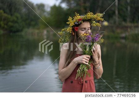Young girl with wreath on her head holds bouquet of wildflowers on river background. Ivan Kupala holiday 77469156