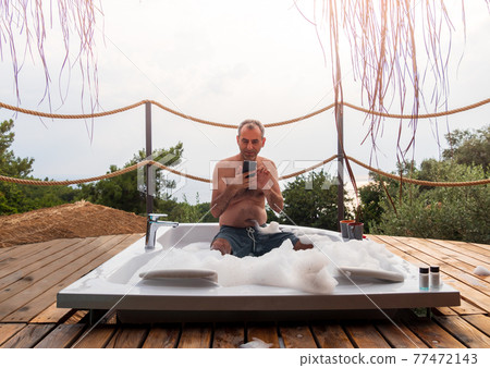 A man looking his smartphone in a hot tub at a luxurious exotic spa resort hotel balcony with sea and sunset view. 77472143