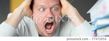 Dumbfounded man holds his head and looks at the stack of folders 77472658
