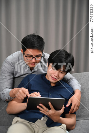 Happy gay couple embracing and using digital on sofa. Homosexual, LGBT concept. Happy gay couple embracing and using digital on sofa. Homosexual, LGBT concept. 77478339