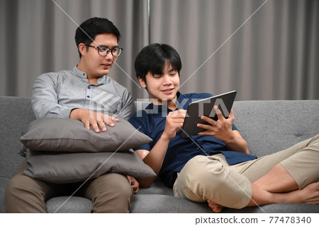 Casual homosexual couple using digital while sitting on sofa at home. Homosexual, LGBT concept. Casual homosexual couple using digital while sitting on sofa at home. Homosexual, LGBT concept. 77478340