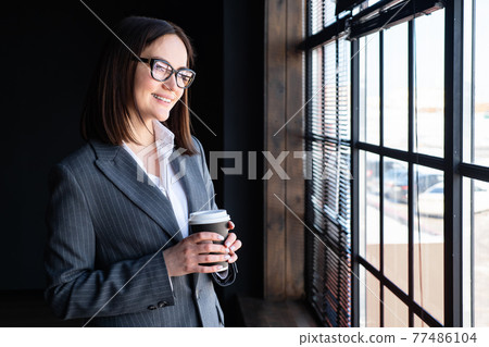 Young woman in a business suit with a drink in a paper glass looks out the window Young woman in a business suit with a drink in a paper glass looks out the window 77486104