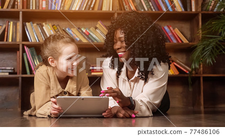 Schoolgirl and babysitter look at tablet display and laugh Schoolgirl and babysitter look at tablet display and laugh 77486106