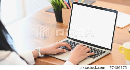 Cropped image of young western woman hands typing on white blank screen computer laptop that putting on wooden desk and surrounded by potted plant and wooden pencil holder. 77486573