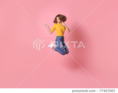 cheerful positive girl jumping in the air with raised hand pointing to copy space isolated on pink background. 77487005