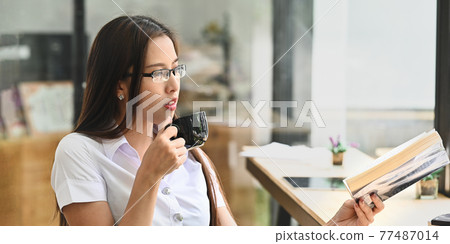 Brunette woman in glasses holding a coffee cup while relaxing/reading a book at the long wooden counter bar over comfortable restaurant as background. Brunette woman in glasses holding a coffee cup while relaxing/reading a book at the long wooden counter bar over comfortable restaurant as background. 77487014