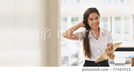 Photo of beautiful smiling woman in white shirt holding a clipboard while standing with keep hand on head over the classroom glass wall as background. Photo of beautiful smiling woman in white shirt holding a clipboard while standing with keep hand on head over the classroom glass wall as background. 77487015