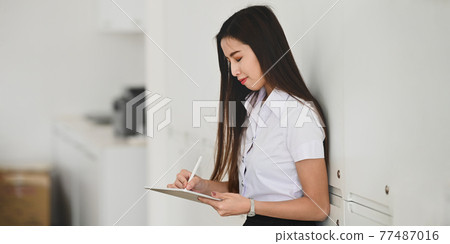Beautiful university student girl drawing on computer tablet by using a stylus pen while standing with lean on lockers over locker room as background. Beautiful university student girl drawing on computer tablet by using a stylus pen while standing with lean on lockers over locker room as background. 77487016