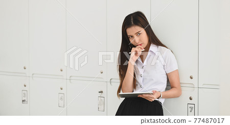 Beautiful university student girl keep hand on chin while standing and holding a computer tablet with stylus pen over locker room as background. Beautiful university student girl keep hand on chin while standing and holding a computer tablet with stylus pen over locker room as background. 77487017