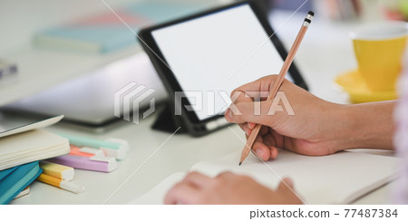 Cropped image of young student hands writing on notebook while sitting in front of white blank screen computer tablet that putting on white student desk. Student doing homework at home concept. 77487384