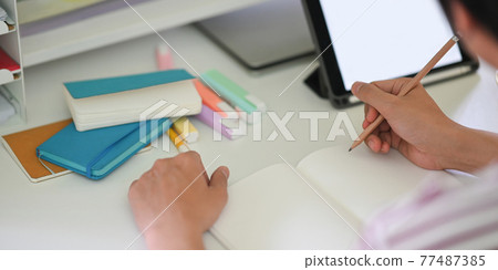 Cropped image of young student hands writing on notebook while sitting in front of white blank screen computer tablet that putting on white student desk. Student doing homework at home concept. 77487385
