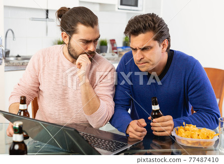 Friendly male meeting over beer at home, men looking at laptop and discussing Friendly male meeting over beer at home, men looking at laptop and discussing 77487410