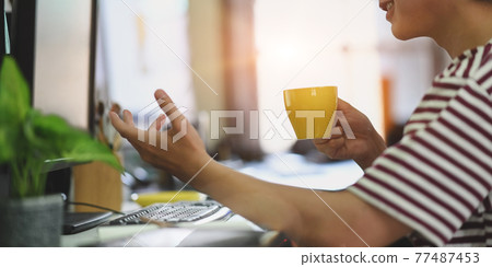 Cropped image of smart man in striped t-shirt drinking a hot coffee while sitting and working with computer at the white working desk in the morning vibe. 77487453