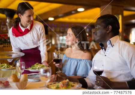 Young waitress bringing dishes to couple at restaurant Young waitress bringing dishes to couple at restaurant 77487576