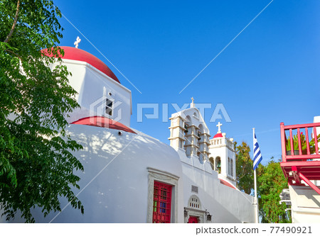 Traditional Greek Orthodox church in Greek island town. Red dome, whitewashed walls, Greek flag and bell tower. Mykonos, Cyclades, Greece. Traditional Greek Orthodox church in Greek island town. Red dome, whitewashed walls, Greek flag and bell tower. Mykonos, Cyclades, Greece. 77490921