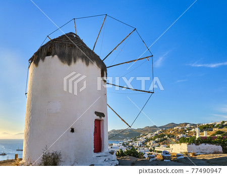 Famous tourist attraction of Mykonos, Greece. Traditional whitewashed windmill on hilltop. Summer, sundown, blue sky. Travel destination, iconic view. Famous tourist attraction of Mykonos, Greece. Traditional whitewashed windmill on hilltop. Summer, sundown, blue sky. Travel destination, iconic view. 77490947