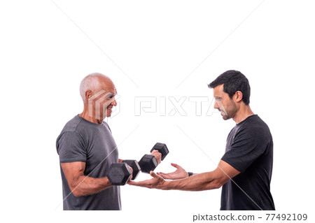 Elderly male client exercising with a fitness trainer, lifts dumbbells. On a white isolated background. 77492109
