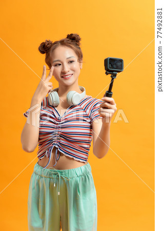 Woman capturing herself with small personal camera on yellow background 77492881
