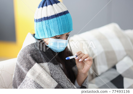 Woman in protective mask and warm hat sitting under covers and looking at thermometer 77493136