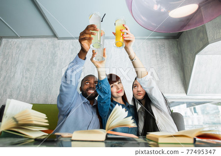 Friendship, youth and food concept. Happy young multiethnic friends, sitting together in cafe with books, drinking fresh juice and celebrating success or passing the exam. Friendship, youth and food concept. Happy young multiethnic friends, sitting together in cafe with books, drinking fresh juice and celebrating success or passing the exam. 77495347